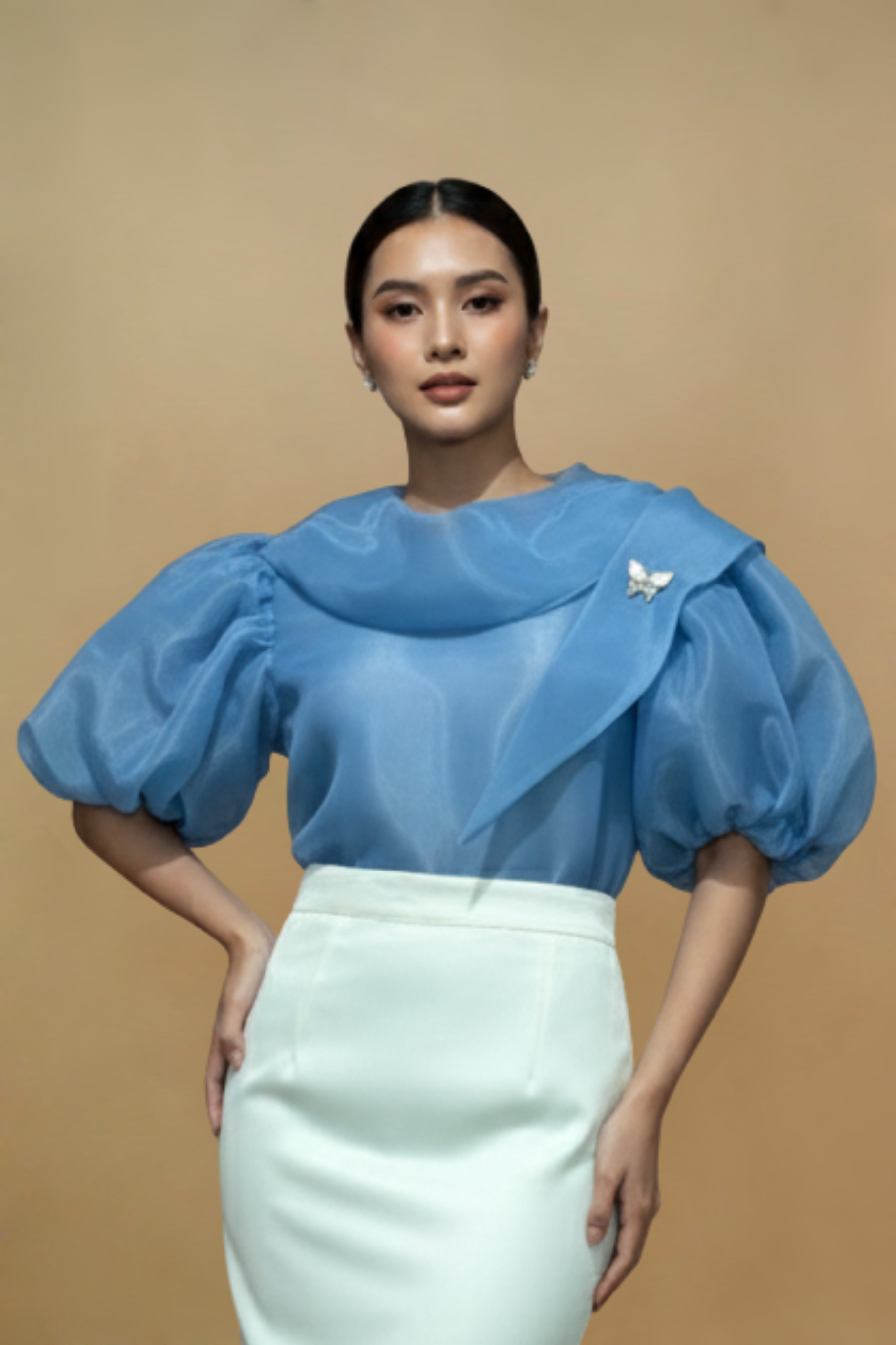 Woman wearing a blue Filipiniana kimona top with puffed sleeves and a white skirt against a beige background